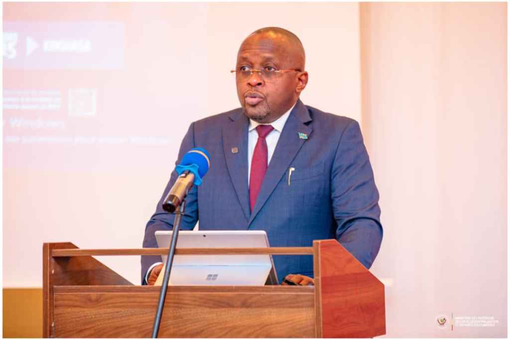 Un homme en costume, debout derrière un pupitre, prononçant un discours lors d'une conférence sur la gouvernance et la prévention de l'extrémisme violent en République Démocratique du Congo.