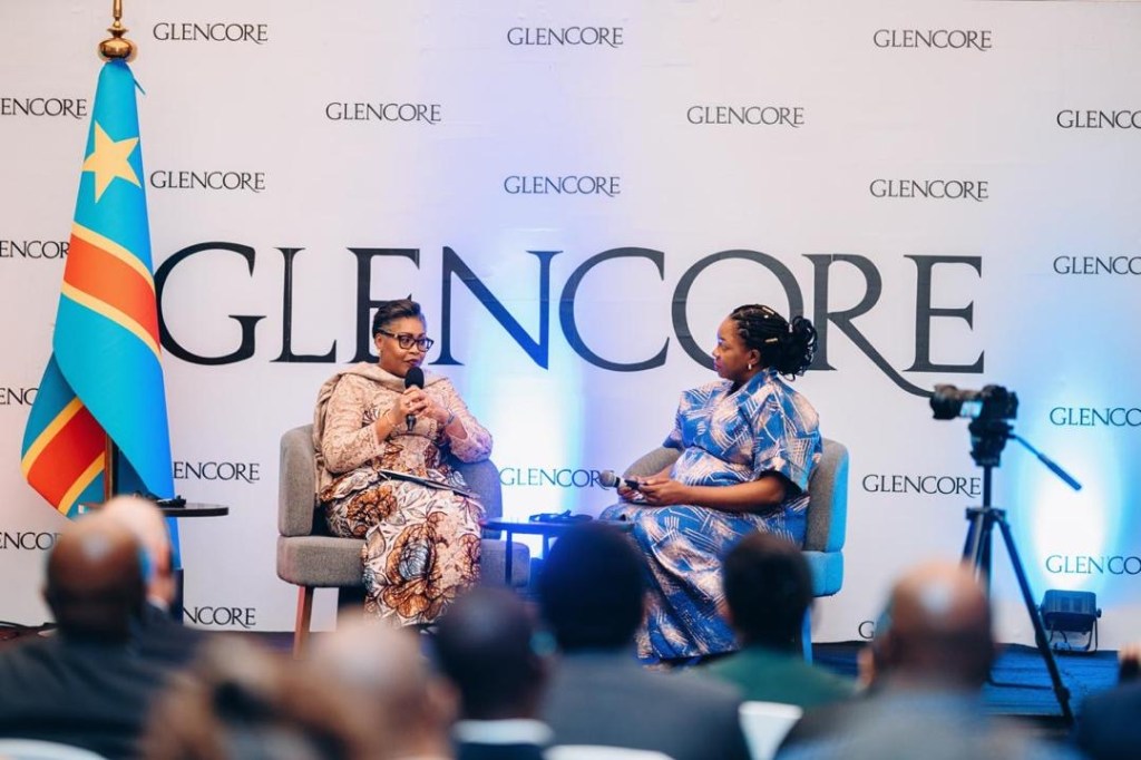 La Première Ministre Judith Suminwa Tuluka en discussion lors de la 7e Conférence annuelle de Glencore, avec un drapeau de la RDC en arrière-plan et le logo de Glencore sur la scène.