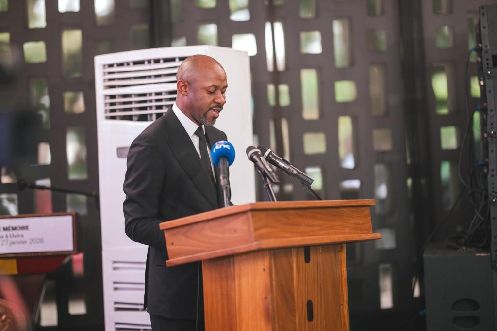 A man in formal attire speaking at a wooden podium with microphones during an event, with a modern backdrop and an air conditioning unit visible in the background.