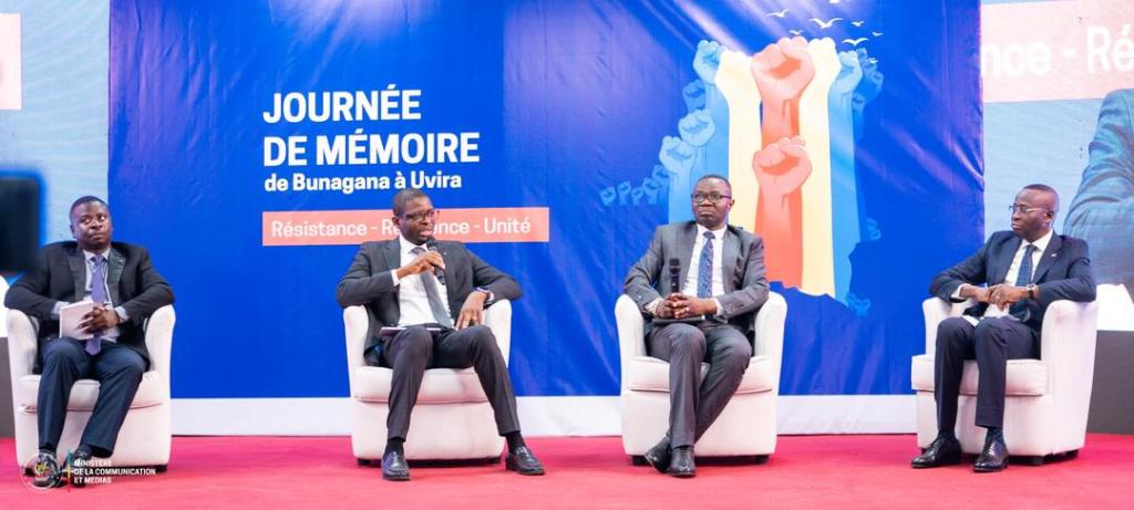 A panel discussion featuring four speakers seated on chairs, set against a blue backdrop with the text 'JOURNÉE DE MÉMOIRE de Bunagana à Uvira' and images of raised fists symbolizing resistance and unity.