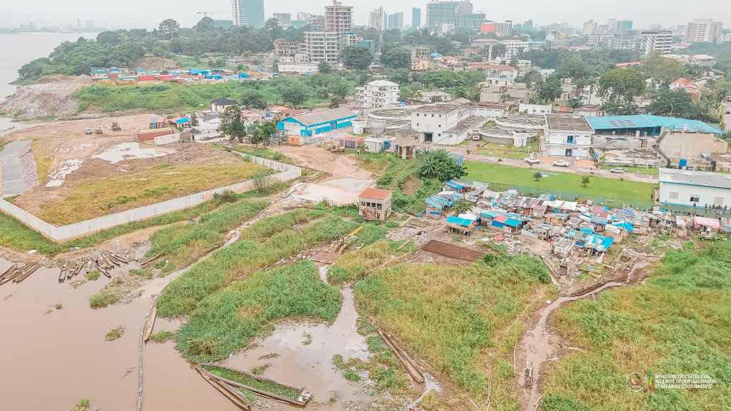 Aerial view of a mixed-use urban area featuring a riverfront with buildings, greenery, and a community of structures with blue rooftops.