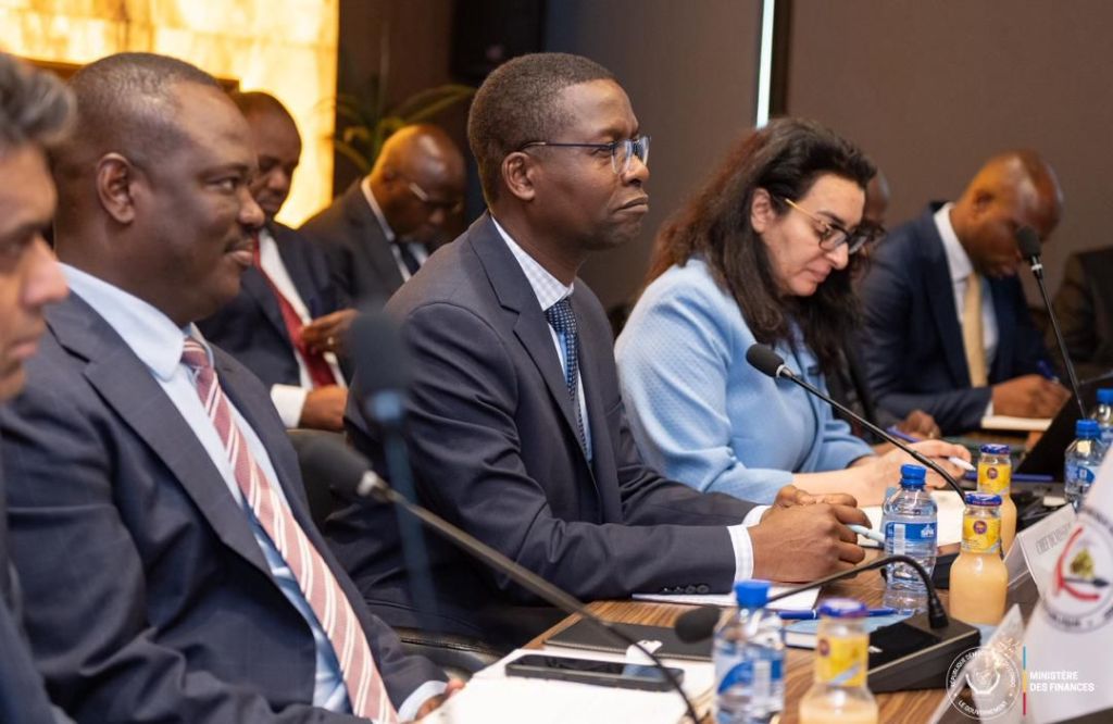 A group of professionals seated at a conference table, engaged in a meeting. They are focused on discussions, with water bottles and notebooks in front of them. The setting appears formal and includes a mix of genders and ethnic backgrounds.